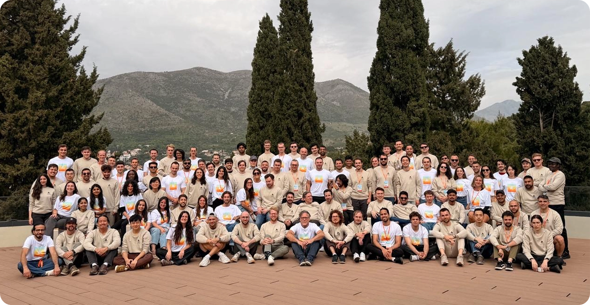 Large diverse group of people wearing matching beige and white team shirts posed together outdoors with mountains and cypress trees in background.