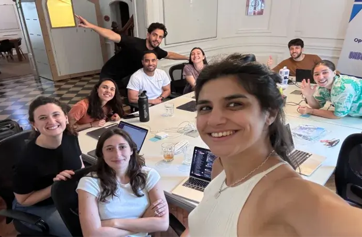 A diverse team of young professionals smiling and posing together around a table in a collaborative workspace.