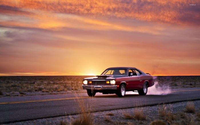 Red vintage car driving on desert highway at sunset with dramatic orange sky.