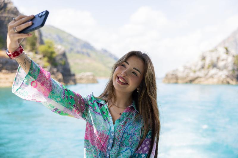 A young woman takes a selfie on a boat with a scenic backdrop of mountains and water. She is wearing a colorful patterned shirt and smiling at the camera.