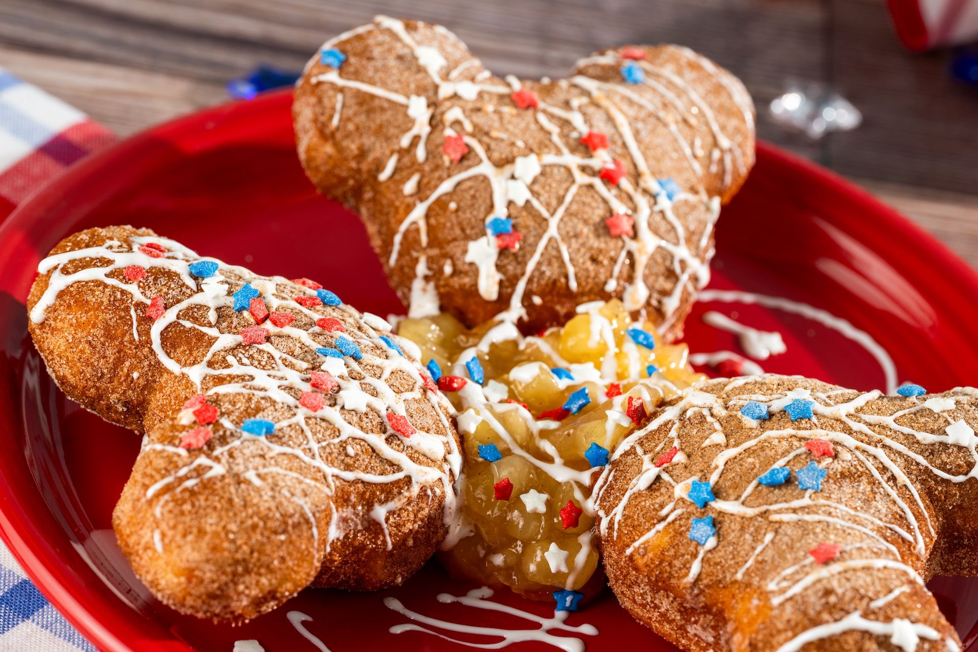 The image features three brown, star-shaped pastries adorned with white icing and red, white, and blue sprinkles. The pastries are arranged on a vibrant red plate, highlighting a festive theme suitable for celebrations.