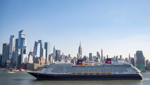 The image captures a large cruise ship navigating through the water near a city skyline, featuring prominent skyscrapers and the Empire State Building in the background. The scene conveys a vibrant urban environment juxtaposed with maritime activity.