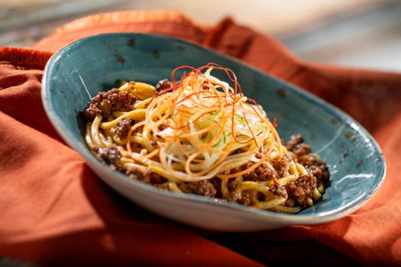 The image showcases a serving of zhajiang noodles, a popular noodle dish, presented in a blue bowl. It features a savory meat topping, garnished with thinly sliced vegetables on a wooden surface.