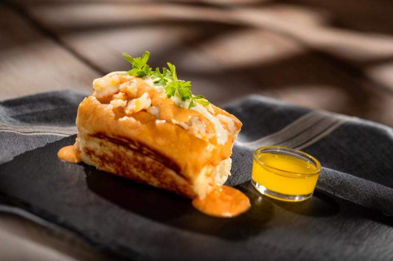 The image features a gourmet seafood dish showcased on a slate plate. It includes a roll topped with shrimp and a drizzle of sauce, accompanied by a small portion of sauce in a separate dish.