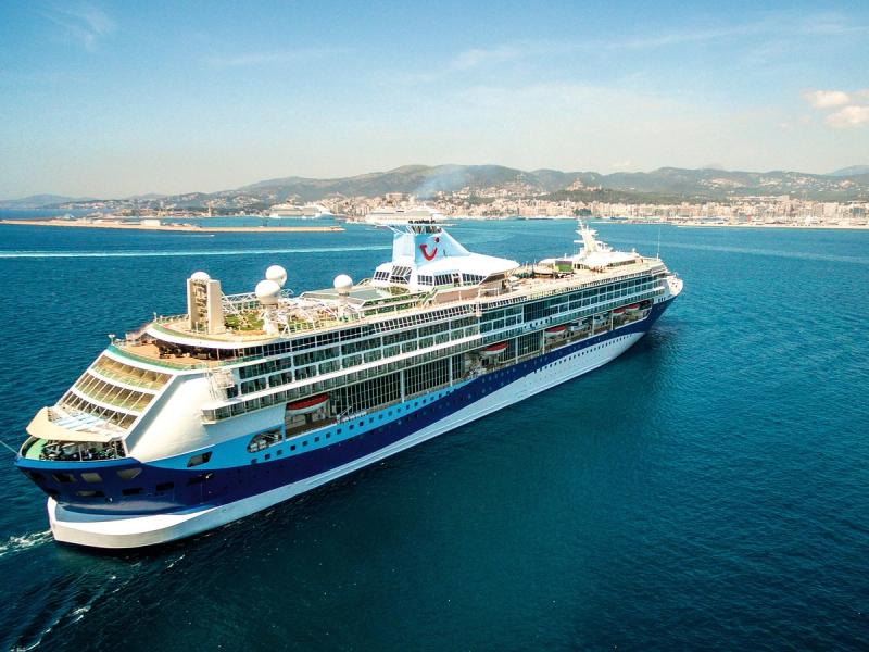 A large cruise ship is sailing through clear blue waters, with a coastline visible in the background. The vessel is equipped with various deck amenities and appears to be on a leisure voyage.