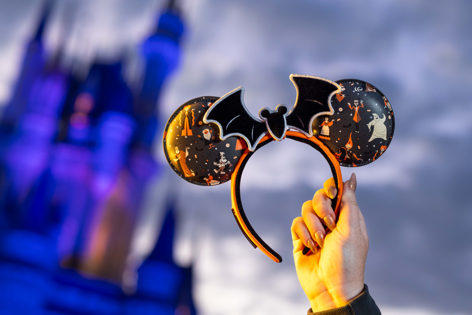 A hand holds a pair of Halloween-themed Mickey Mouse ears featuring a bat design in front of a castle backdrop. The image showcases a festive atmosphere, highlighting the playful elements of a celebration at a theme park.