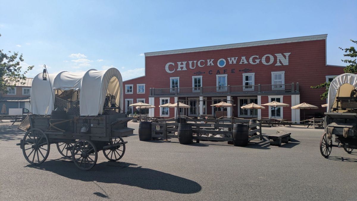 The image depicts a vintage-style cafe named 'Chuck Wagon Cafe' set against a clear blue sky. In the foreground, there are two covered wagons positioned on a pavement, suggesting an outdoor dining area with tables and umbrellas.