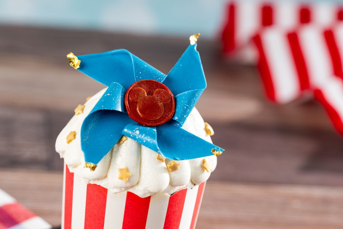 A close-up of a decorative cupcake featuring a blue pinwheel design and a red circular decoration with a Mickey Mouse silhouette. The cupcake is presented in a striped paper liner, creating a festive appearance.