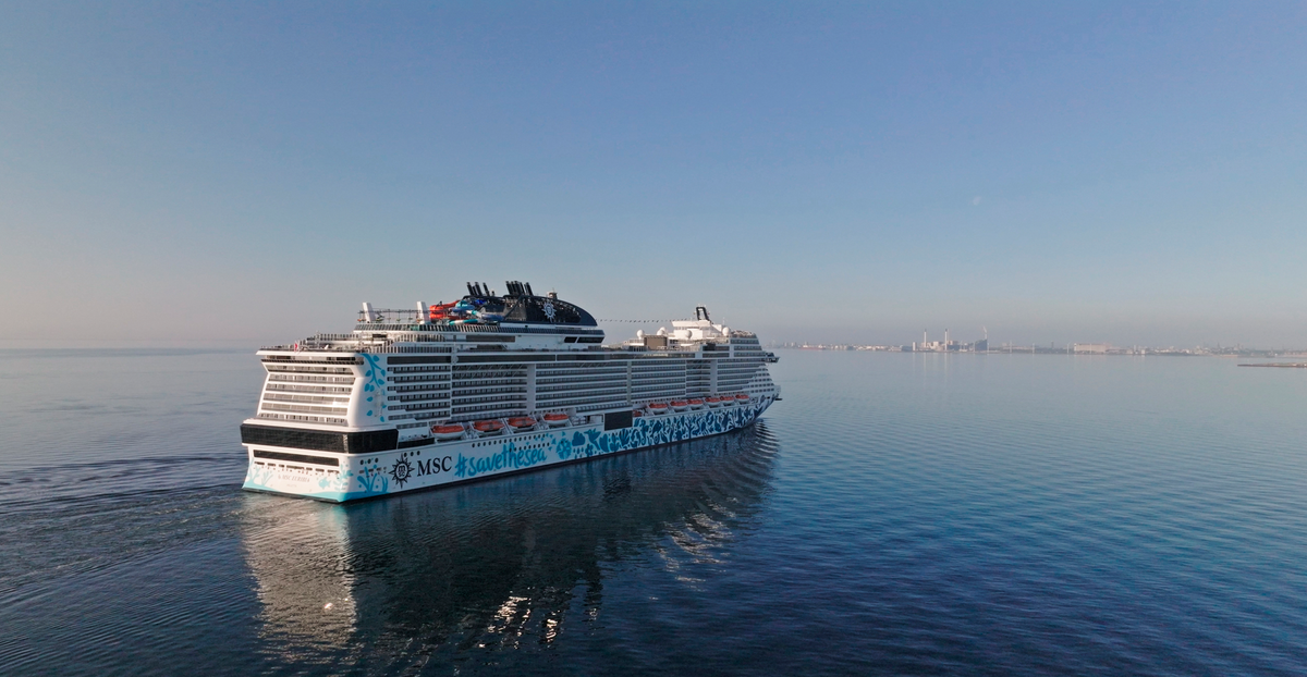 The image shows the MSC Euribia, a large cruise ship, sailing on calm waters with a clear blue sky in the background. The ship features a colorful design on its hull, reflecting in the water as it moves toward the horizon.