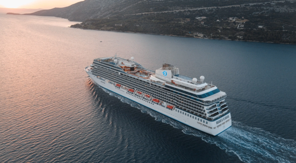 The image shows a large cruise ship navigating through calm waters during sunset, with a mountainous coastline in the background. The ship features multiple decks and amenities for passengers.