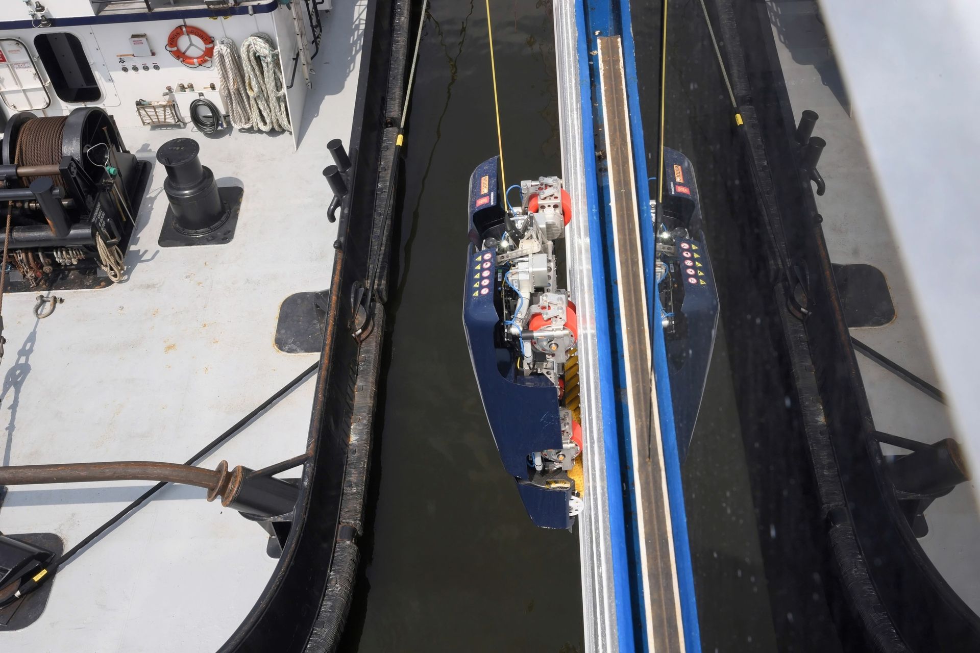 The image shows the mechanical apparatus of a winch positioned above the water, flanked by the sides of a vessel. The view is from above, capturing the details of the winch as it operates near the water's edge.