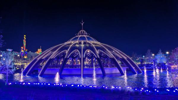The image features a beautifully illuminated structure resembling a futuristic dome, surrounded by a water feature that includes light displays. The scene is set at night, highlighting the vibrant colors and intricate design of the architecture.