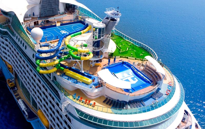 The image depicts the upper deck of a large cruise ship featuring various recreational amenities. There are water slides, a golf putting green, and sports courts visible, all situated above the ocean.