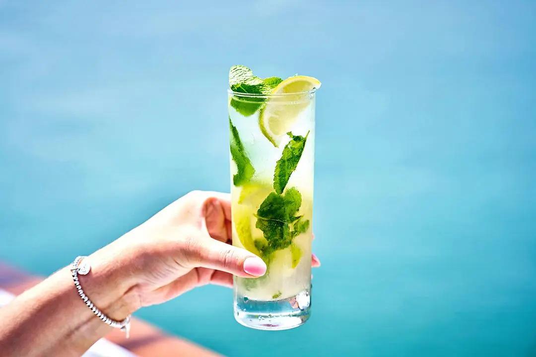 A hand holding a tall glass of mojito cocktail, filled with fresh mint leaves, lime slices, and ice. The background features a serene blue water setting, suggesting a tropical or beach atmosphere.