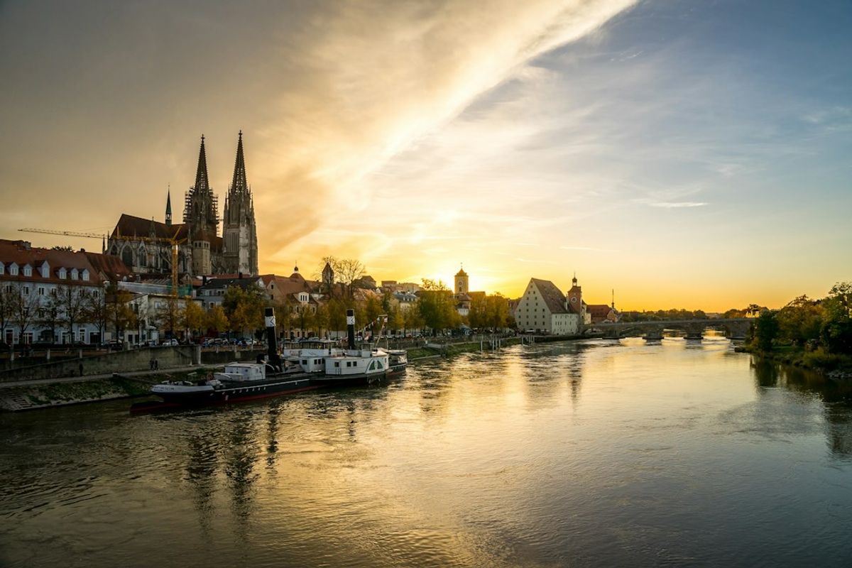 The image captures a scenic view of a riverside city at sunset, featuring historic architecture and a calm water surface reflecting the sky's colors. The silhouette of a prominent church with tall spires dominates the skyline.