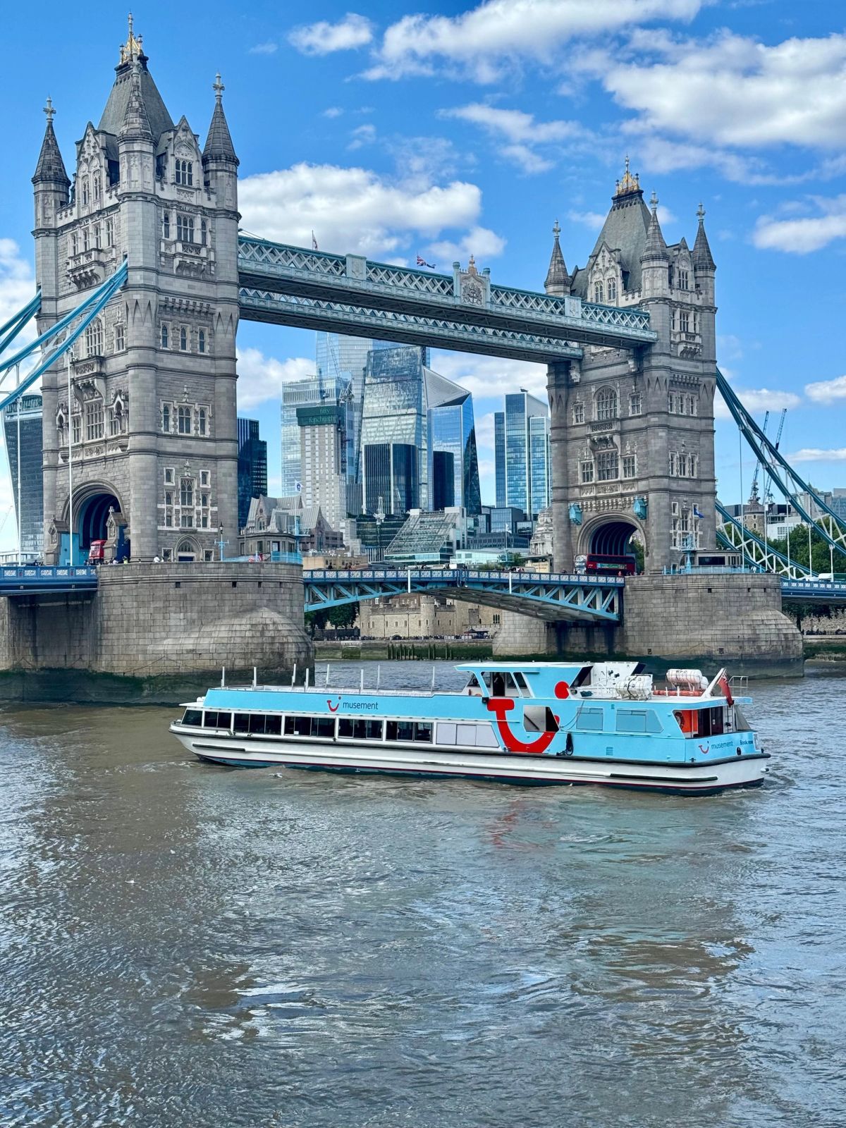 A modern boat navigates the River Thames in London, passing under the iconic Tower Bridge. In the background, the city skyline showcases a blend of historic and contemporary architecture.