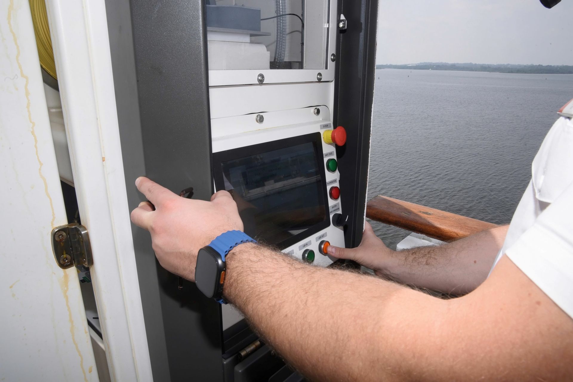 An individual is operating a control panel aboard a vessel near a body of water. The focus is on the person's hands adjusting the controls, highlighting the technology involved in navigation or operation.