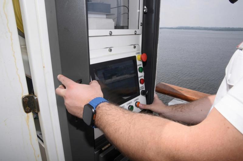 An individual is operating a control panel aboard a vessel near a body of water. The focus is on the person's hands adjusting the controls, highlighting the technology involved in navigation or operation.