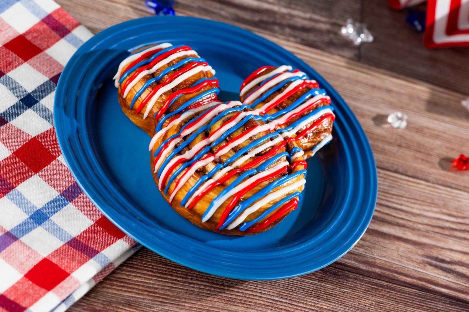 A patriotic-themed dessert shaped like a Mickey Mouse head, decorated with red, white, and blue icing. The dessert is placed on a blue plate with a checkered tablecloth and festive decorations in the background.