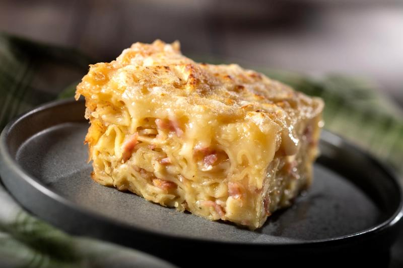 A close-up view of a slice of schinkennudeln, a German dish made with noodles and ham, baked until golden and crispy. The slice is presented on a dark plate, highlighting its creamy texture and cheesy topping.
