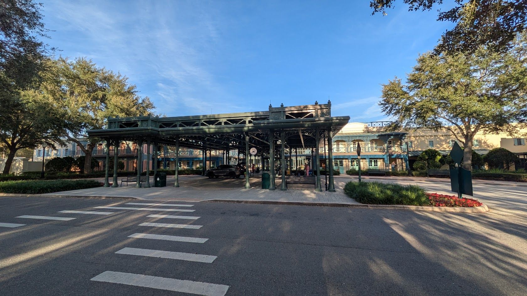 The image depicts a historic structure with a covered platform supported by green iron columns, surrounded by trees and landscaping. In the background, there is a building with large windows, suggesting a blend of old and new architecture.