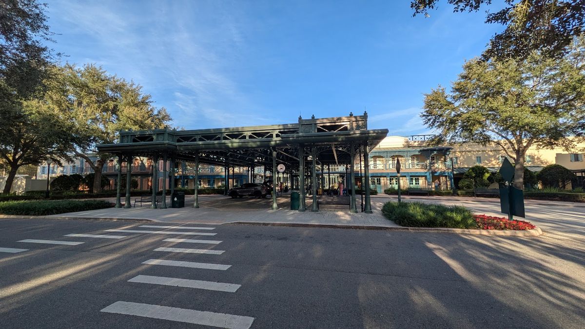 The image depicts a historic structure with a covered platform supported by green iron columns, surrounded by trees and landscaping. In the background, there is a building with large windows, suggesting a blend of old and new architecture.