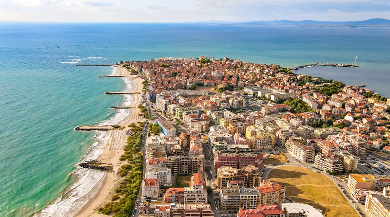 This aerial image showcases a coastal town featuring a blend of residential buildings and sandy beaches along the shoreline. The landscape includes a harbor area and a clear view of the blue ocean in the background, highlighting the town's proximity to water.