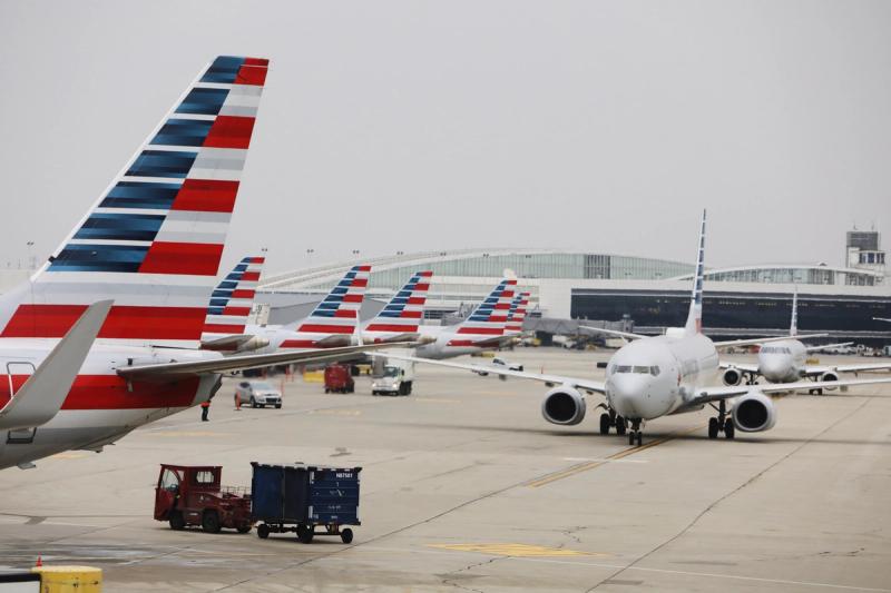 The image features multiple American Airlines aircraft on the tarmac at an airport. The planes, prominently displayed with their distinctive tail designs, are parked or taxiing at the airport, creating a busy atmosphere.