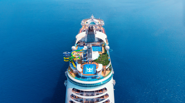 An aerial view of a large cruise ship sailing on calm blue waters. The ship features various recreational facilities including water slides, pools, and green areas on its deck.