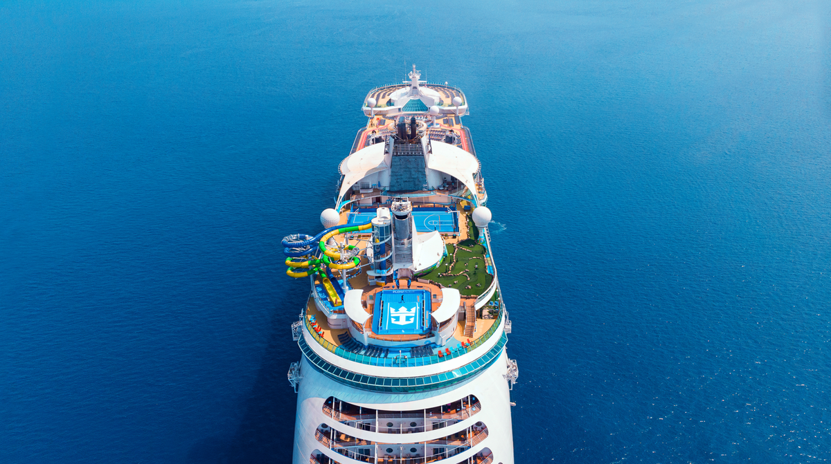 An aerial view of a large cruise ship sailing on calm blue waters. The ship features various recreational facilities including water slides, pools, and green areas on its deck.