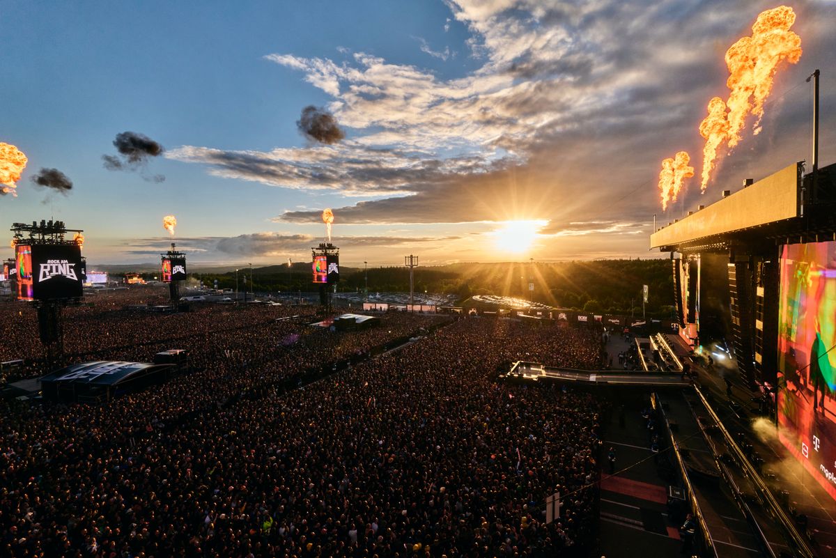 The image captures a large crowd at an outdoor music festival during sunset, with vibrant colors in the sky. Fireworks or flame jets are visible, enhancing the celebratory atmosphere of the event.