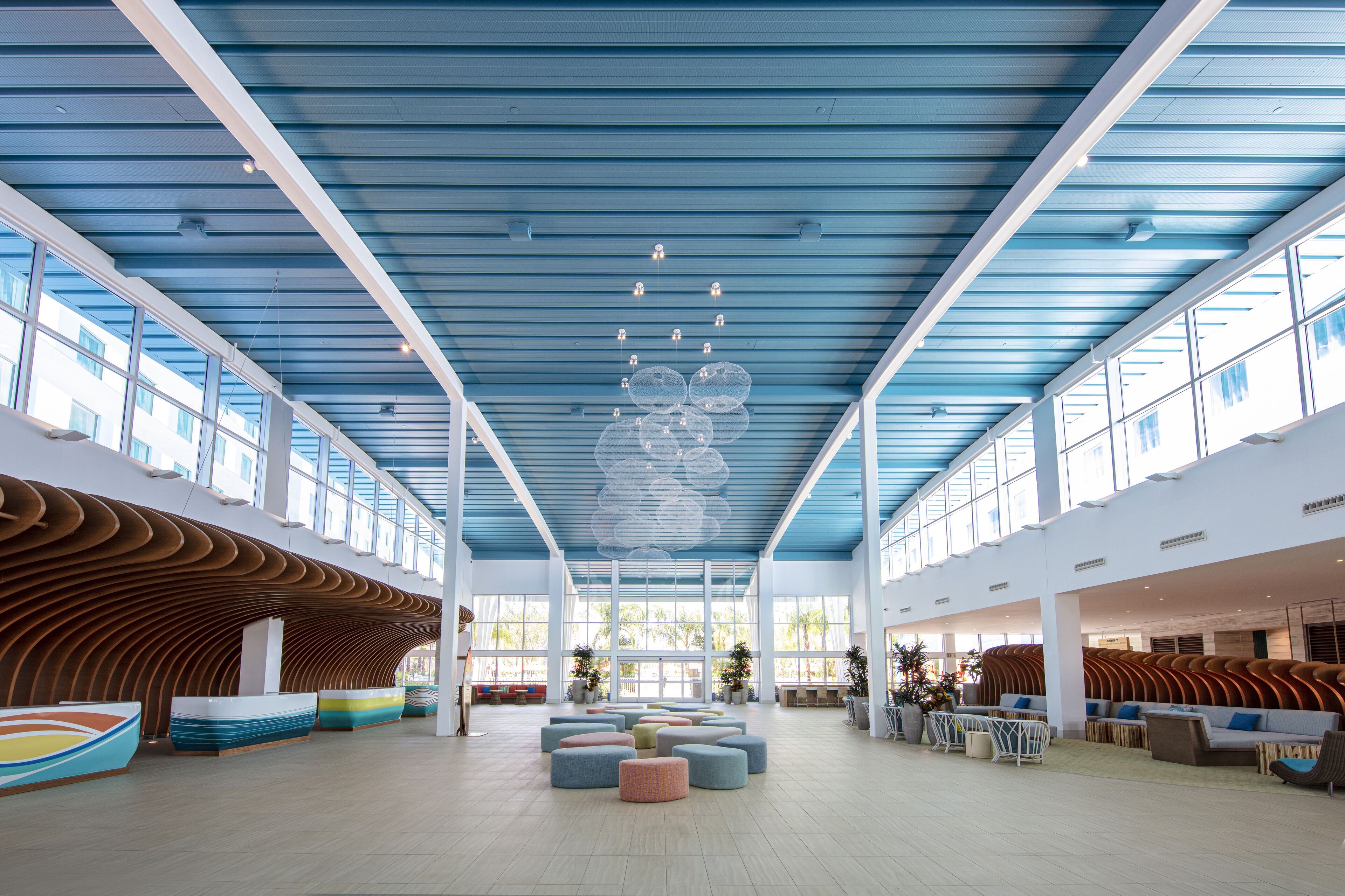 The image showcases a modern interior of a spacious lobby, featuring a unique ceiling design with blue panels and elegant lighting. The space includes a mix of seating arrangements with colorful ottomans and wooden accents, creating a welcoming atmosphere.