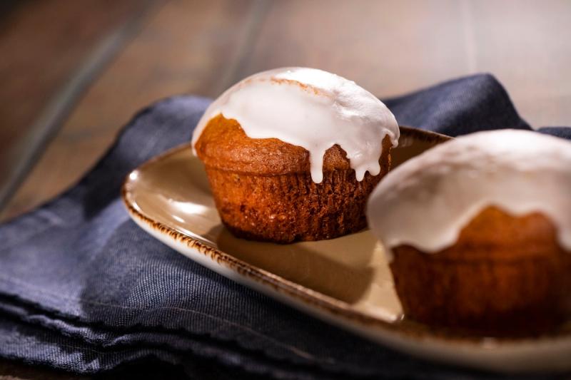 The image features two freshly baked muffins topped with a glossy white icing. They are arranged on a beige plate, placed on a dark blue cloth backdrop, highlighting their texture and details.