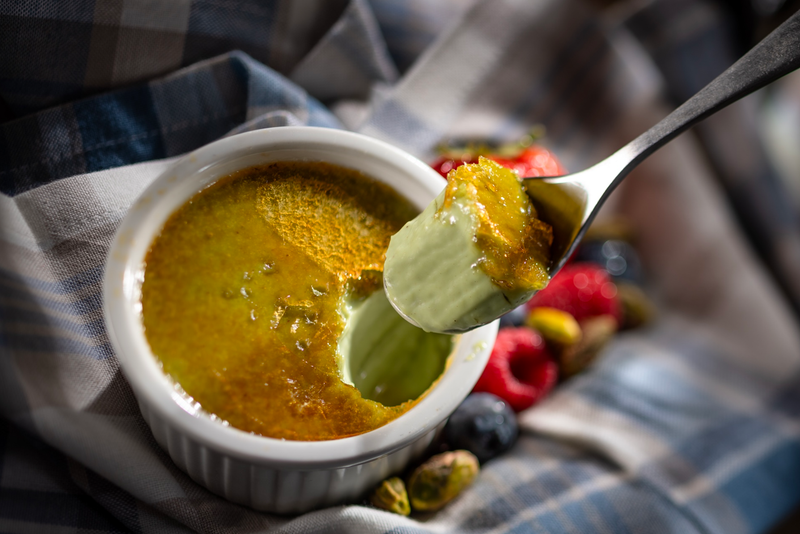 The image showcases a serving of crème brûlée in a small white ramekin. A spoonful of the rich dessert highlights its creamy texture and caramelized sugar topping, surrounded by fresh berries and nuts.