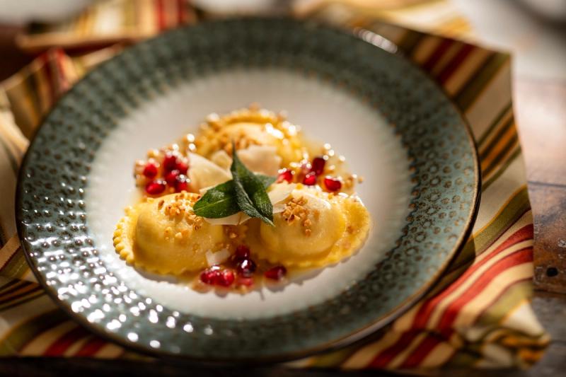 A beautifully plated dish featuring three pieces of ravioli, garnished with pomegranate seeds and a leaf of mint. The dish is presented on a decorative plate, surrounded by a striped cloth backdrop, emphasizing the culinary art of fine dining.