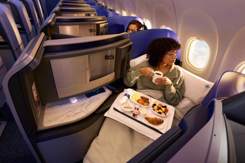 The image depicts a passenger in a premium airline seat enjoying a meal while traveling on an airplane. The passenger is seated beside a window, which showcases a view outside, and is seen holding a cup alongside a tray filled with food items.