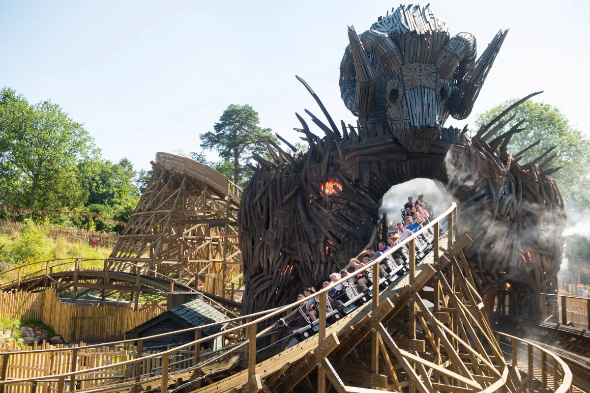 An elaborate wooden structure resembling a giant creature serves as the entrance to a roller coaster at a theme park. Visitors are seen boarding the ride, with some fog effects enhancing the atmosphere.