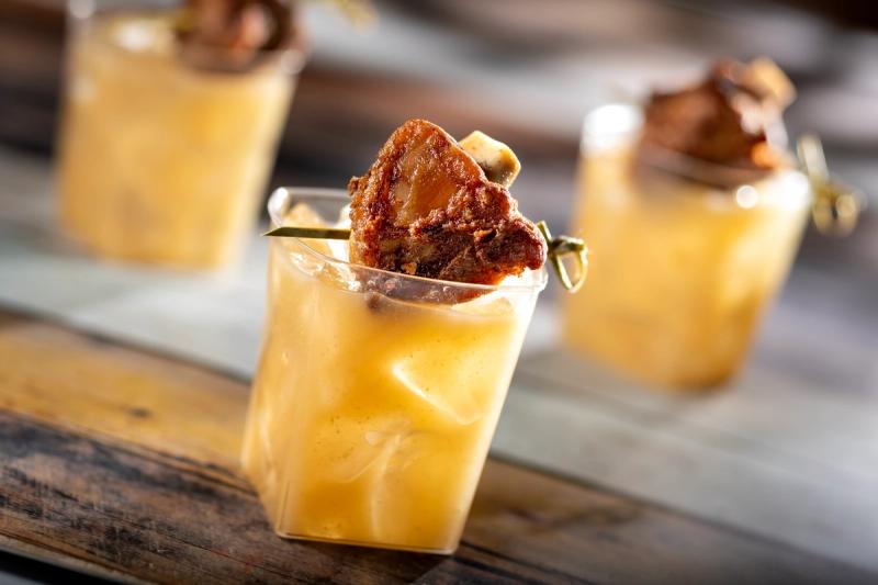 The image features a close-up of a cocktail served in a clear glass, garnished with a piece of crispy meat on a skewer. The background consists of a wooden surface, adding a rustic feel to the presentation.