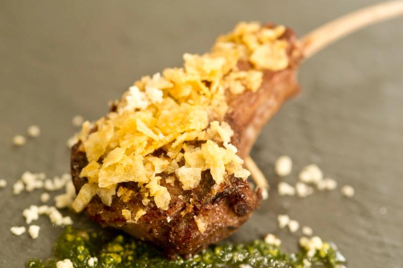 The image features a gourmet lamb chop garnished with crispy potato flakes and drizzled with a vibrant green sauce. The dish is presented on a textured gray surface, highlighting the rich colors and textures of the food.