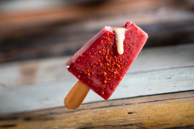 The image showcases a vibrant red paleta, a type of Mexican frozen treat, with a bite taken out revealing a creamy filling. The paleta is set against a rustic wooden background, emphasizing its colorful appearance.