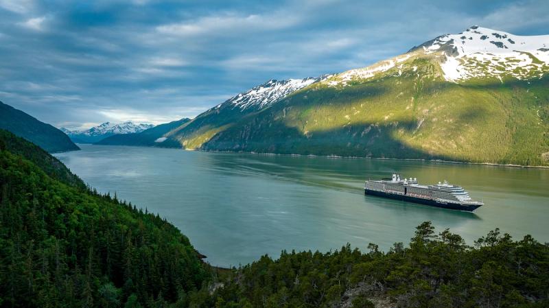 A large cruise ship is anchored in a serene fjord surrounded by lush green forests and snow-capped mountains. The tranquil water reflects the clouds and mountains, creating a picturesque landscape.