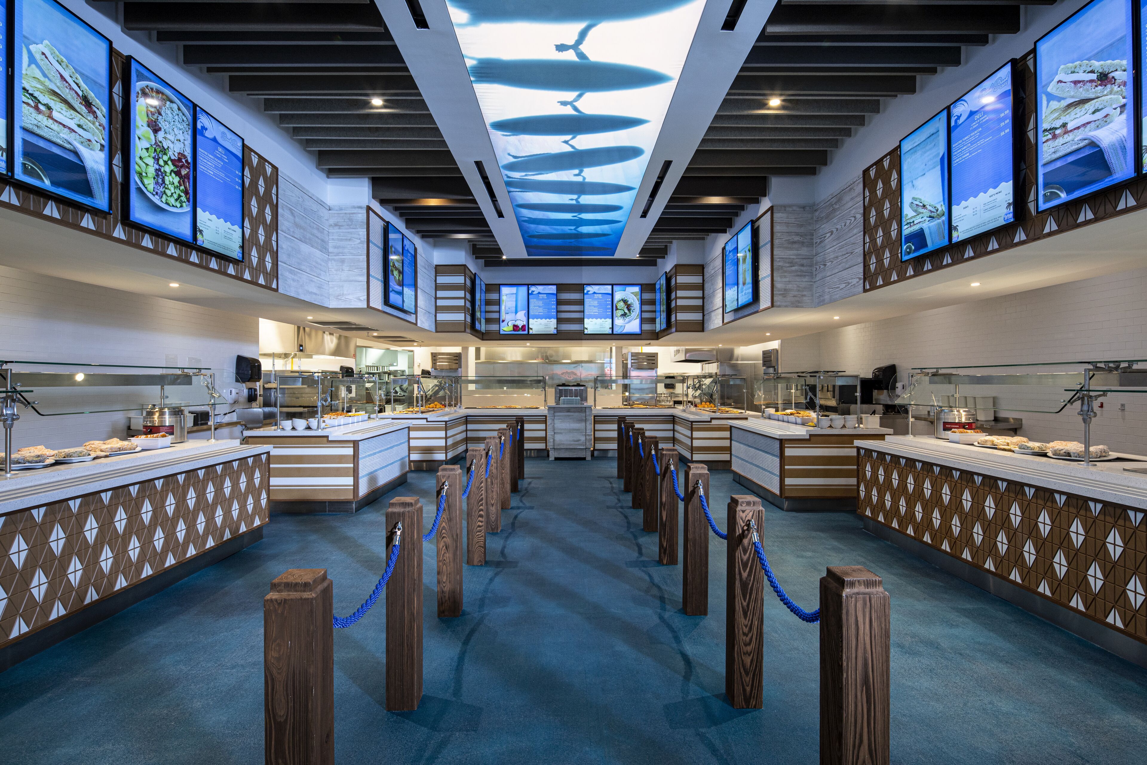 The image depicts a modern dining area featuring a clean, open layout with multiple food stations. Large screens display seafood dishes along the walls, while an aquarium-themed ceiling adds a unique ambiance.