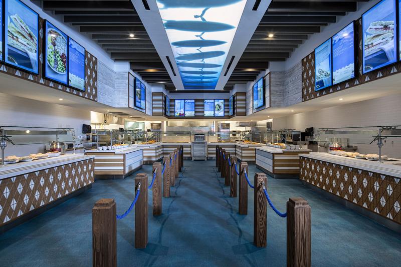 The image depicts a modern dining area featuring a clean, open layout with multiple food stations. Large screens display seafood dishes along the walls, while an aquarium-themed ceiling adds a unique ambiance.