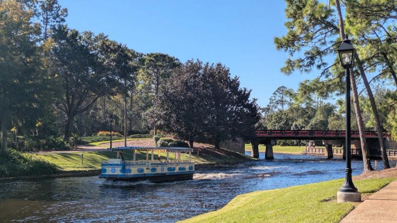 A scenic view of a canal with a small boat cruising along the water. In the background, a pedestrian bridge crosses over the waterway, surrounded by lush greenery and trees under a clear blue sky.