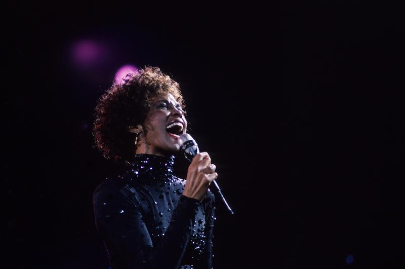 A singer passionately performs on stage, illuminated by soft lights. The artist is captured mid-performance, showcasing an emotional and powerful expression.