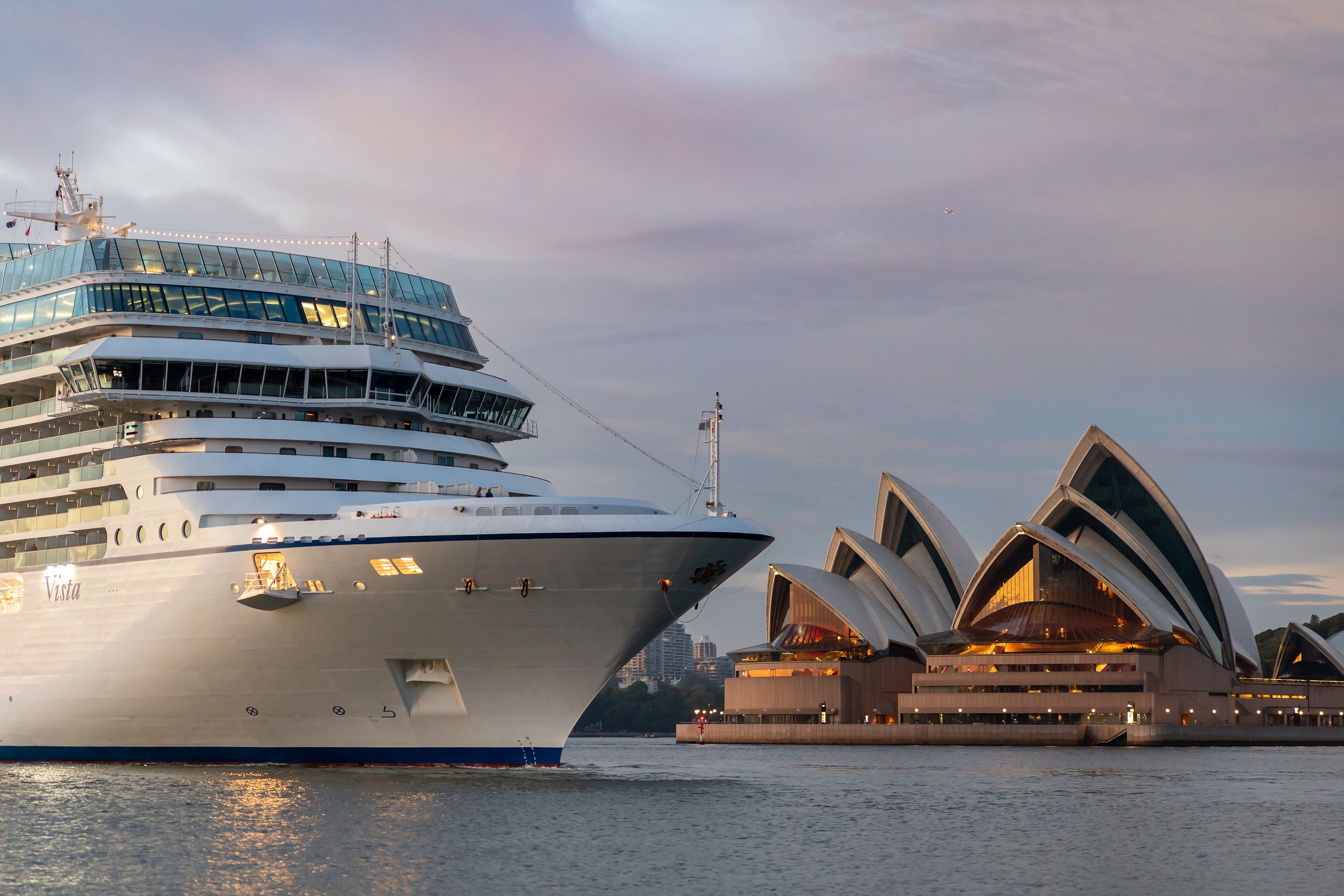 A large cruise ship navigates the waters near the iconic Sydney Opera House at sunset. The ship, with its sleek design, contrasts with the architectural beauty of the opera house, highlighting both maritime travel and cultural landmarks.