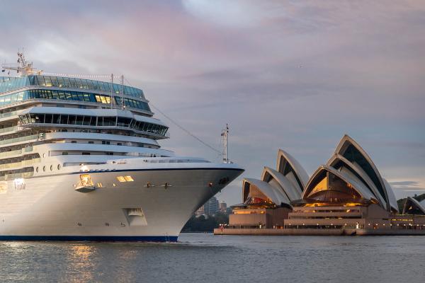 A large cruise ship navigates the waters near the iconic Sydney Opera House at sunset. The ship, with its sleek design, contrasts with the architectural beauty of the opera house, highlighting both maritime travel and cultural landmarks.