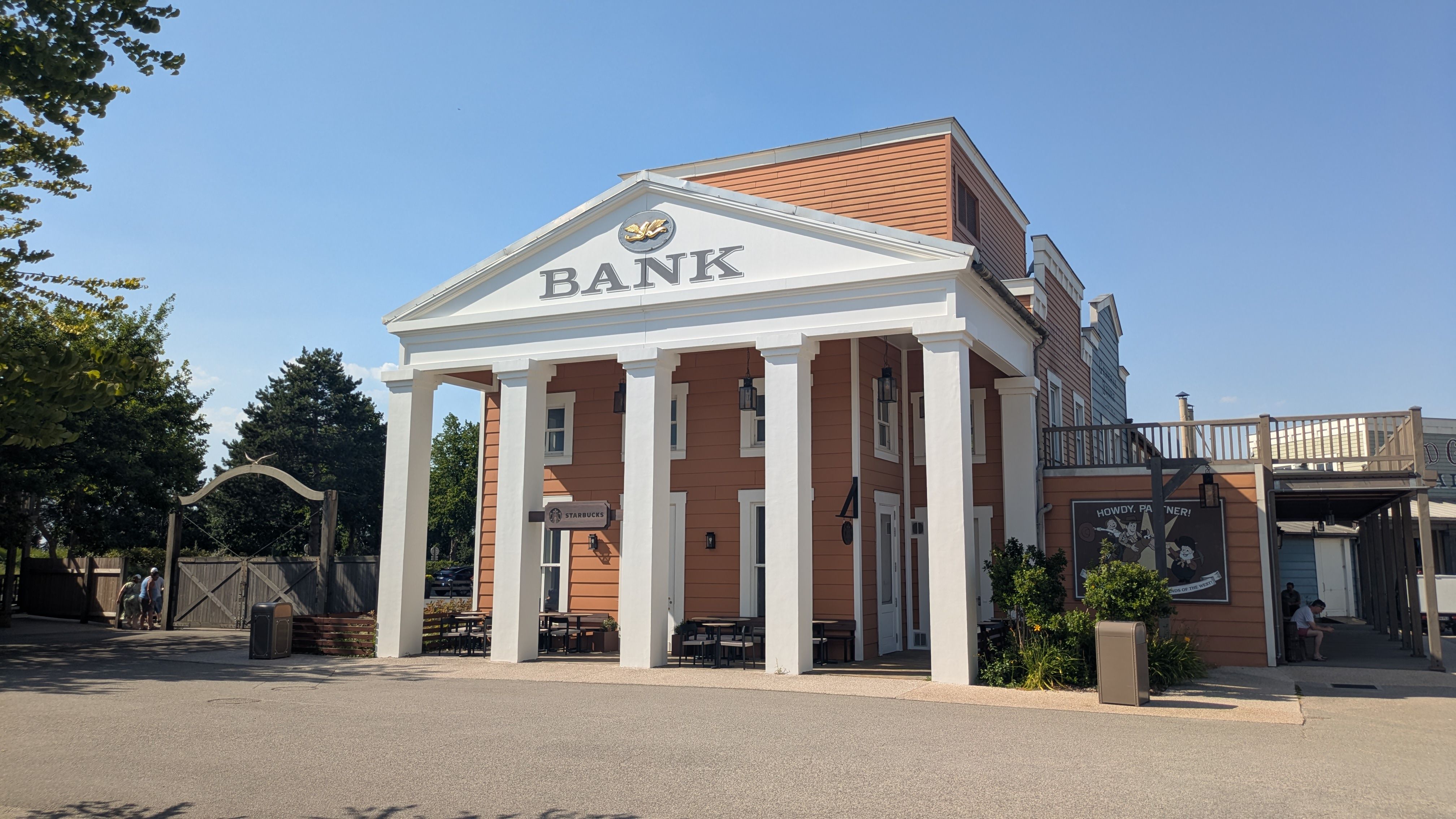 The image depicts a large building with a prominent 'BANK' sign on its facade, featuring traditional architectural elements like white columns. The structure is surrounded by landscaping and includes outdoor seating.