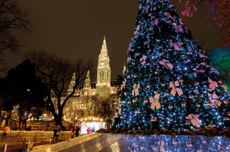 A beautifully decorated Christmas tree adorned with lights and ornaments stands prominently in the foreground, with a historic building illuminated in the background. The night scene captures the festive atmosphere of a holiday celebration in an urban setting.