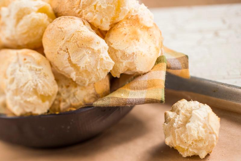 A close-up image of a pile of golden-brown bread rolls, displayed in a black bowl lined with a checkered cloth. One roll is placed beside the bowl, highlighting the texture and freshness of the baked goods.
